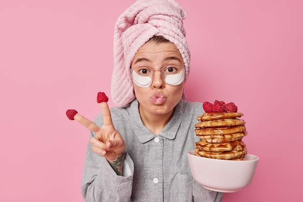 Funny young woman makes peace gesture keeps lips folded poses with delicious homemade pancakes applies beauty patches under eyes dressed in nightwear wears wrapped towel on head poses indoor