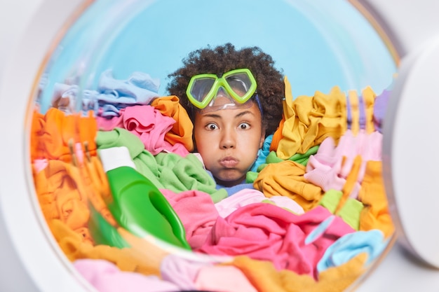 Funny young woman with curly hair wears snorkeling mask on forehead blows cheeks poses around multicolored laundry blue wall