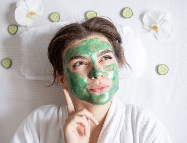 Funny young woman with a green cosmetic mask on her face is resting while lying in the spa salon, top view.