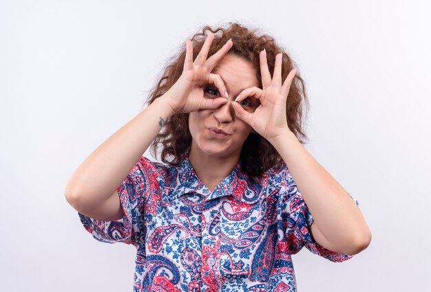 Funny young woman with short curly hair  in colorful shirt doing ok signs with fingers like binocularr looking through fingers 