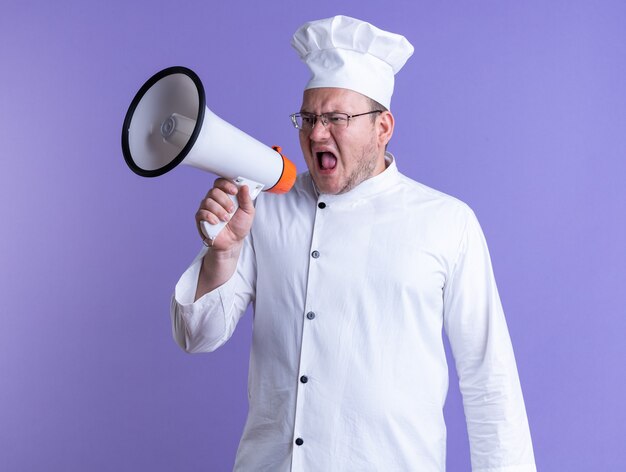 Furioso maschio adulto cuoco indossando l'uniforme dello chef e occhiali guardando a lato gridando in altoparlante isolato su parete viola