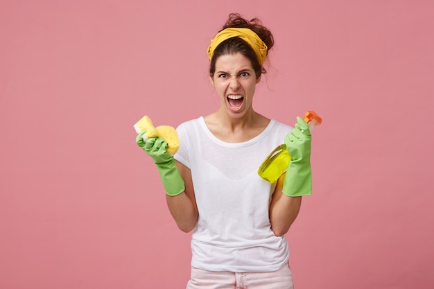 Furious and annoyed woman with yellow scarf on head and in green gloves holding washing spay and sponge having angry look while going to have spring cleaning. Chores, housekeeping and tidying up