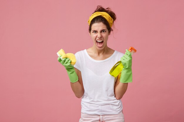 Разъяренная и раздраженная женщина с желтым шарфом на голове и в зеленых перчатках держит спрей для стирки и губку с сердитым взглядом, собираясь на генеральную уборку. Работа по дому, уборка и уборка