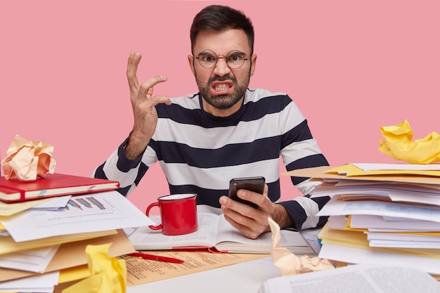 Foto gratuita furioso giovane ragazzo barbuto gesticola con la mano, arrabbiato per ricevere spam sul cellulare, indossa occhiali, maglione a righe bianche e nere