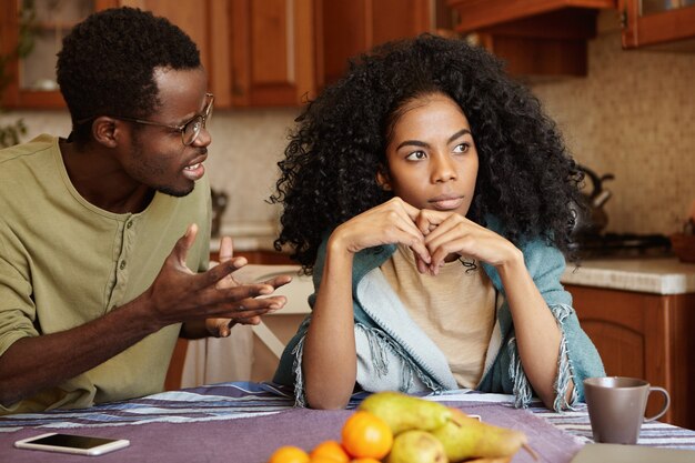 Furious black male gesturing in despair or anger while trying to make excuses to his offended wife as if saying: Can you just hear me out? African couple going through hard time in relationships
