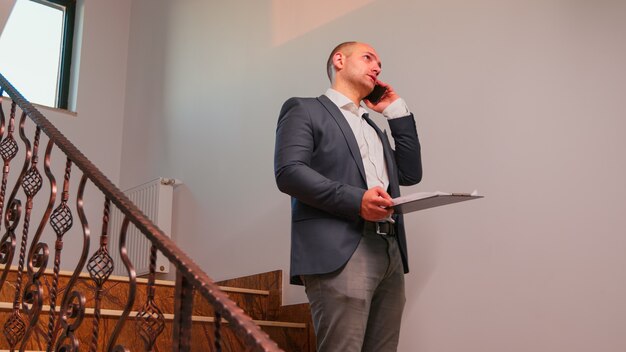 Furious businessman talking on smartphone standing on stairs in finance corporate company working overtime. Group of professional successful businesspeople working in modern finacial building.