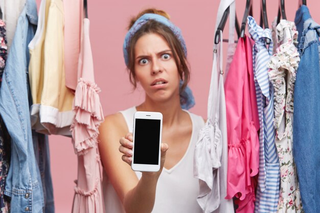 Furious female with exhausted look, standing between clothes, showing mobile phone while standing in cloakroom. Young female with discontent expression doing shopping, using smart phone indoors