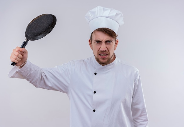 흰 벽에 프라이팬으로 공격하는 흰색 밥솥 유니폼과 모자를 쓰고 분노 잘 생긴 젊은 수염 난 요리사 남자