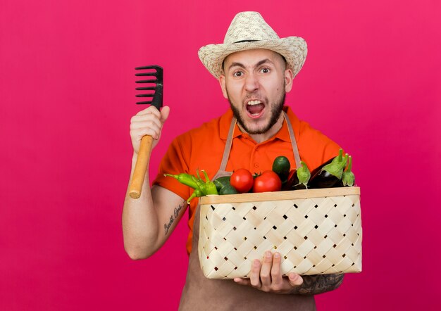 Разъяренный садовник в садовой шляпе держит корзину с овощами и грабли