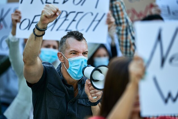 Furious man wearing protective face mask shouting through megaphone while protesting with crowd of people during coronavirus pandemic