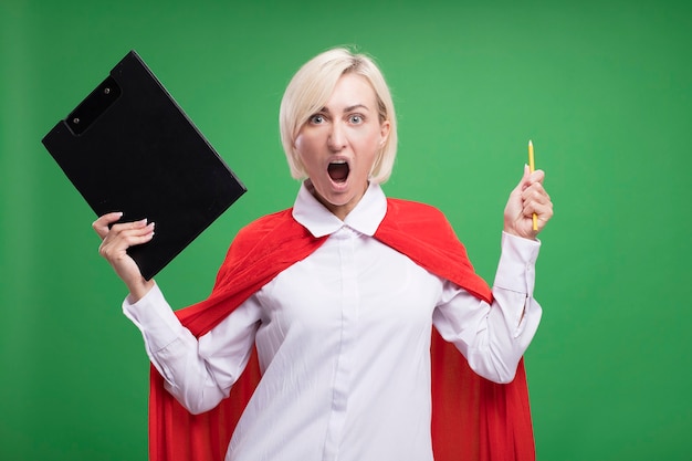 Free photo furious middle-aged blonde superhero woman in red cape holding clipboard and pencil looking at front screaming isolated on green wall