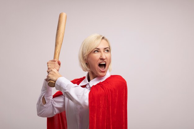 Furious middle-aged blonde superhero woman in red cape raising baseball bat looking at side screaming isolated on white wall with copy space