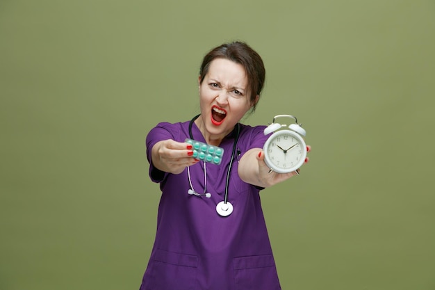 разъяренная женщина-врач средних лет в униформе и со стетоскопом на шее смотрит в камеру, растягивая пачку таблеток и будильник в сторону камеры, крича изолированно на оливково-зеленом фоне
