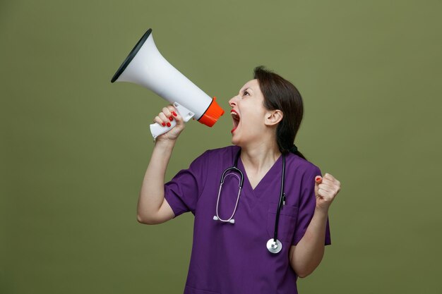 furious middleaged female doctor wearing uniform and stethoscope around neck turning head to side keeping fist in air looking at side shouting out loudly into speaker