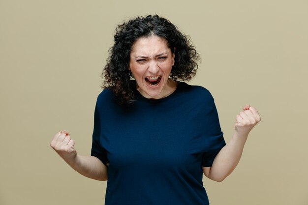 Furious middleaged woman wearing tshirt looking at camera shouting out loudly while keeping fists in air isolated on olive green background