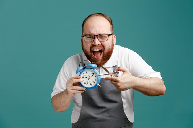 Furious young barber wearing uniform and glasses looking at camera holding alarm clock trying to cut alarm clock with scissors isolated on blue background