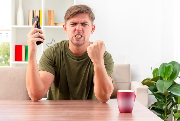 분노한 젊은 금발의 잘 생긴 남자는 컵이 전화를 들고 거실 안에 주먹을 유지하면서 테이블에 앉아있다.