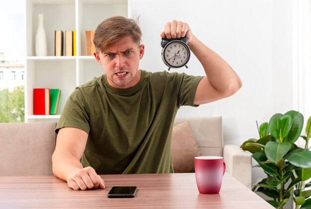 Furious young blonde handsome man sits at table with cup and phone holding alarm clock and looking at camera inside living room