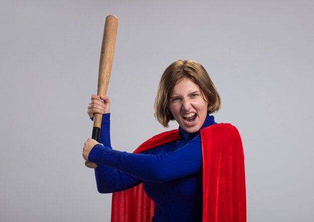 Furious young blonde superhero girl in red cape standing in profile view holding baseball bat  getting ready to hit screaming isolated on white wall with copy space