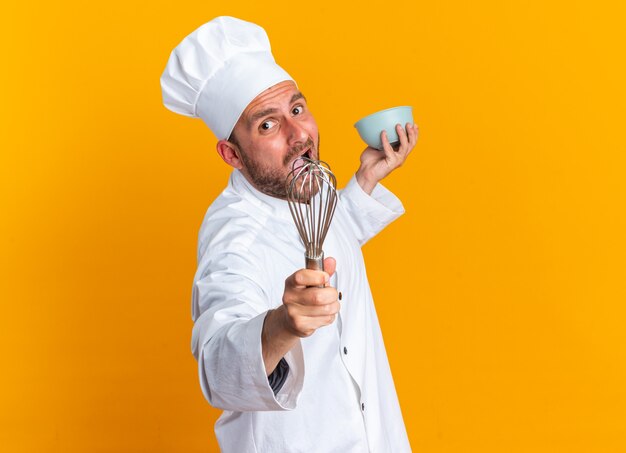 Furious young caucasian male cook in chef uniform and cap standing in profile view looking at camera stretching out whisk and bowl isolated on orange wall with copy space