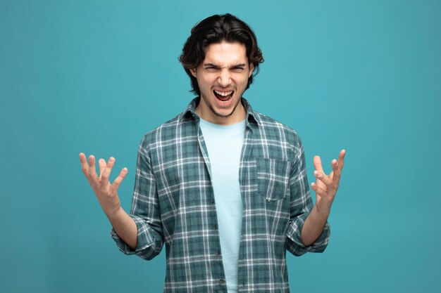 furious young handsome man keeping hands in air looking at camera screaming isolated on blue background