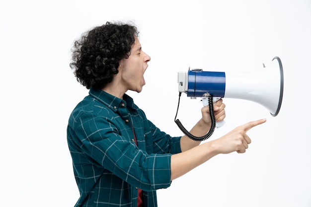 Furious young handsome man standing in profile view pointing to side shouting out loud into speaker with closed eyes isolated on white background