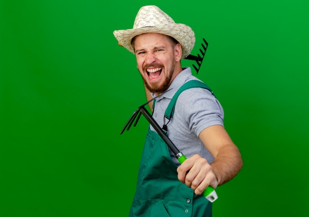 Foto gratuita furioso giovane giardiniere slavo bello in uniforme e cappello cercando di tenere rastrello e zappa-rastrello