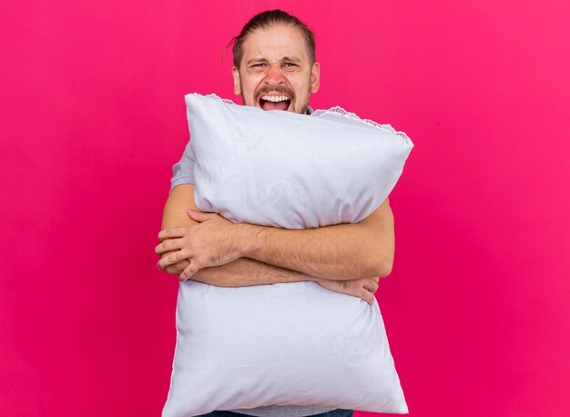 Furious young handsome slavic ill man hugging pillow looking at front screaming isolated on pink wall with copy space