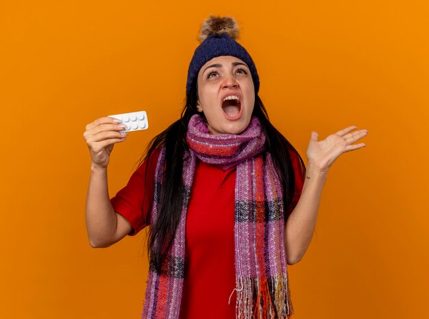 冬の帽子とスカーフを身に着けている猛烈な若い病気の女性は、空の手を見せて、オレンジ色の壁に孤立して叫んで見上げるタブレットのパックを保持しています
