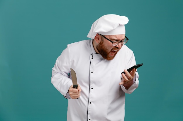 Furioso giovane chef maschio che indossa occhiali uniforme e cappuccio che tiene il coltello che tiene e guarda il telefono cellulare gridando ad alta voce isolato su sfondo blu