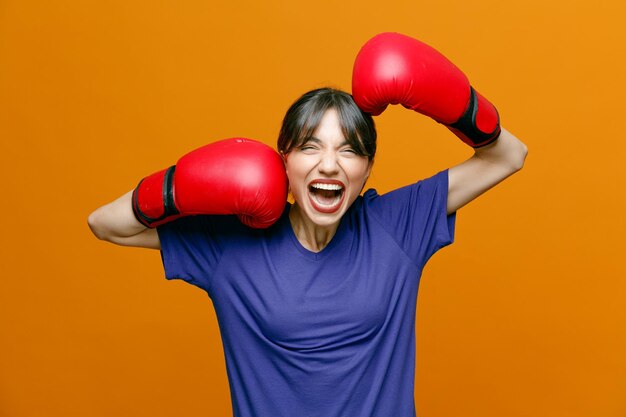 티셔츠를 입고 권투 장갑을 끼고 권투 장갑을 끼고 주황색 배경에 격리된 카메라를 보고 있는 격렬한 젊은 여성