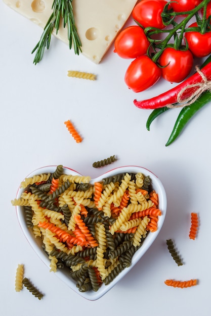 Fusilli pasta with tomatoes, peppers, plant, cheese in a bowl on white table, top view.