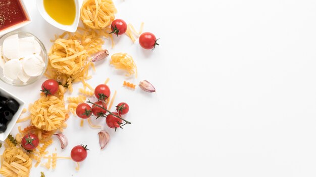 Fusilli and tagliatelle raw pasta with oil; sauce; olives; cheese; tomato and garlic clove above white surface