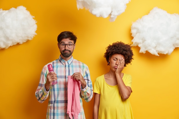 Il futuro padre prepara gli oggetti per il nascituro, tiene il biberon con capezzolo e cursori, la moglie incinta stanca sta vicino, copre il viso con il palmo, si prepara per la nascita del bambino. concetto di genitorialità