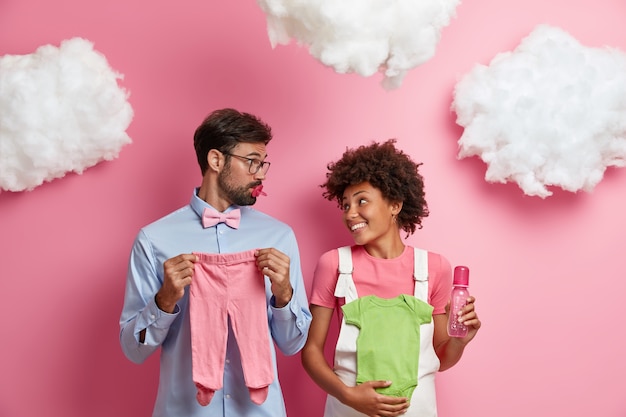 Free photo future happy parents try to guess gender of baby, pose with child sliders, singlet, bottle for feeding and nipple, expect child birth, pose against rosy wall with fluffy white clouds above