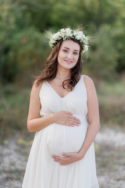 Future mum in white dress and wreath is holding belly on the warm day outdoors