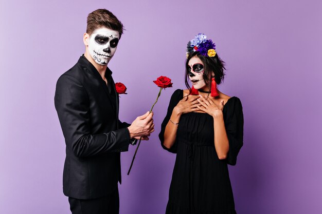 Gallant young man is pleasant to surprise his beloved girl by giving her rose. Shot of Mexican couple with face art in form of skull.