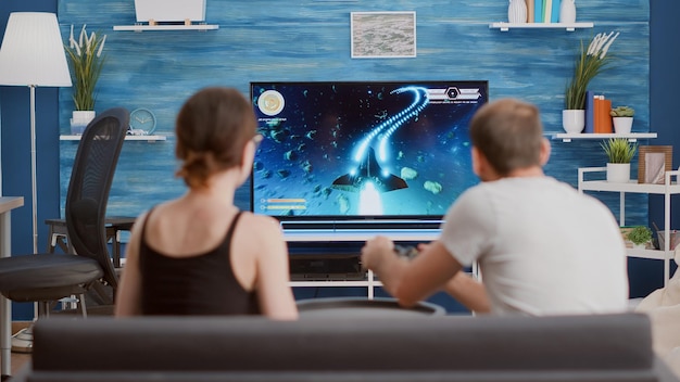 Gamer playing console video game first person shooter on tv while girlfriend is eating popcorn and giving him advice sitting on sofa. Couple on couch enjoying gaming simulation in modern living room.