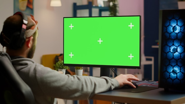 Gamer playing video games on powerful computer with green screen chroma key desktop mock-up display in gaming home studio. Player using RGB keyboard with isolated monitor streaming game wearing headse