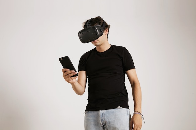 A gamer in VR glasses and plain black t-shirt looking at his smartphone isolated on white