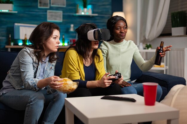 비디오 게임을 하는 조이스틱을 들고 가상 현실 헤드셋을 착용한 게이머 여성과 함께 시간을 보내는 온라인 경쟁에서 그녀를 돕는 동안 다민족 친구들. 수다떨기 개념