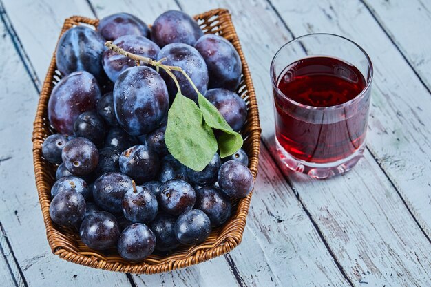 Garden plums in a basket on blue with a glass of juice.