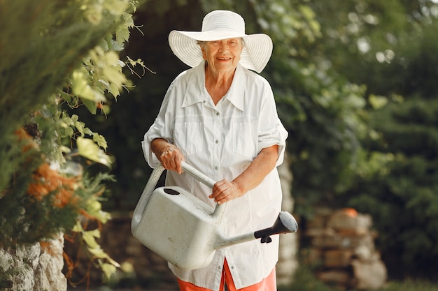 Садоводство летом. Женщина поливает цветы из лейки. Старая женщина в шляпе.