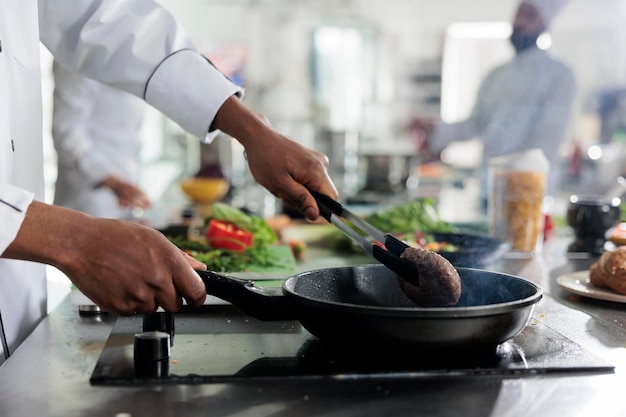 Gastronomy expert cooking succulent beef patty in hot pan while cooking gourmet dish for dinner service at restaurant. Food industry worker preparing meat for meal in professional kitchen.