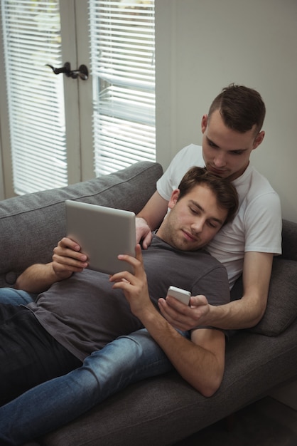 소파에 누워있는 동안 디지털 태블릿 및 휴대 전화를 사용하는 게이 커플
