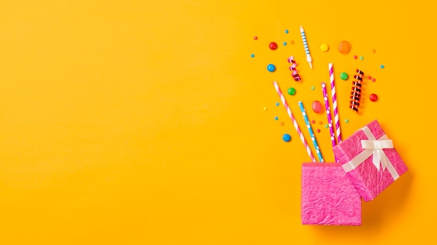 Gems; drinking straws; streamers; sprinkles from the open pink box on yellow backdrop
