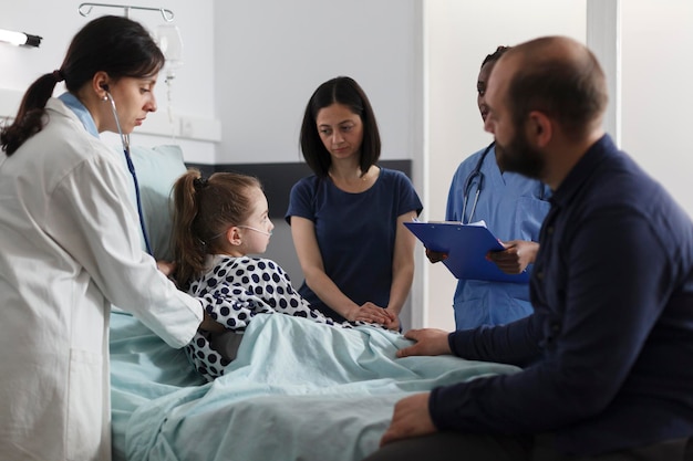 Foto gratuita medico di medicina generale che consulta una bambina malata in cura all'interno del reparto pediatrico della clinica. pediatra esperto che controlla le condizioni di salute della ragazza malata usando lo stetoscopio mentre i genitori si siedono accanto a lei.