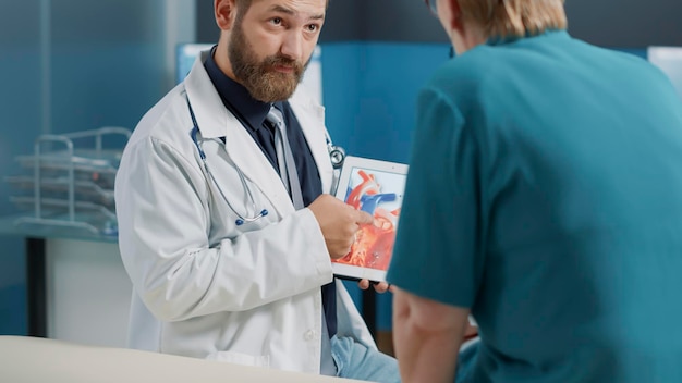 General practitioner holding tablet with heart condition and cardiology illustration at consultation with old patient. Doctor explaining cardiovascular system and diagnosis to elder woman.