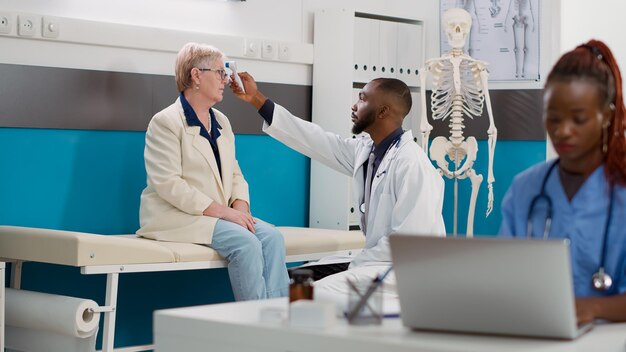 General practitioner using thermometer to check temperature on senior patient at consultation appointment. Doctor doing fever measurement with tool at checkup visit, healthcare support.