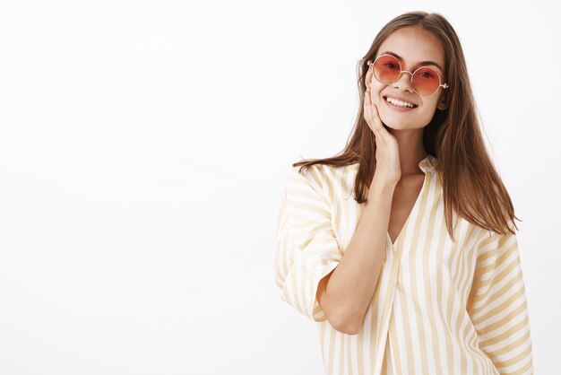 gentle attractive and stylish young sociable woman with brown hair in red sunglasses and white summer blouse touching face and smiling